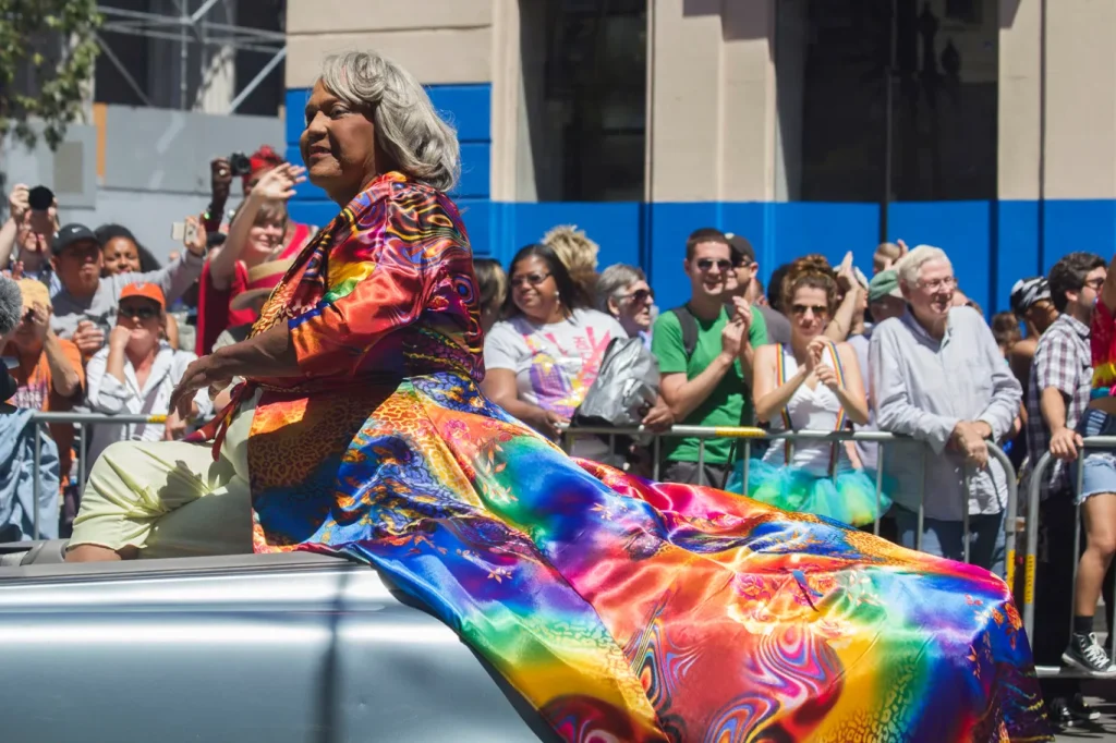 Miss Major riding in a convertible with a rainbow tie dye cloth in a parade